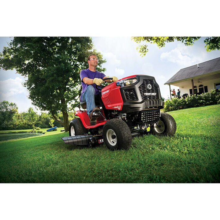 Man operating a red riding lawn mower in a grassy area with trees and a house in the background.